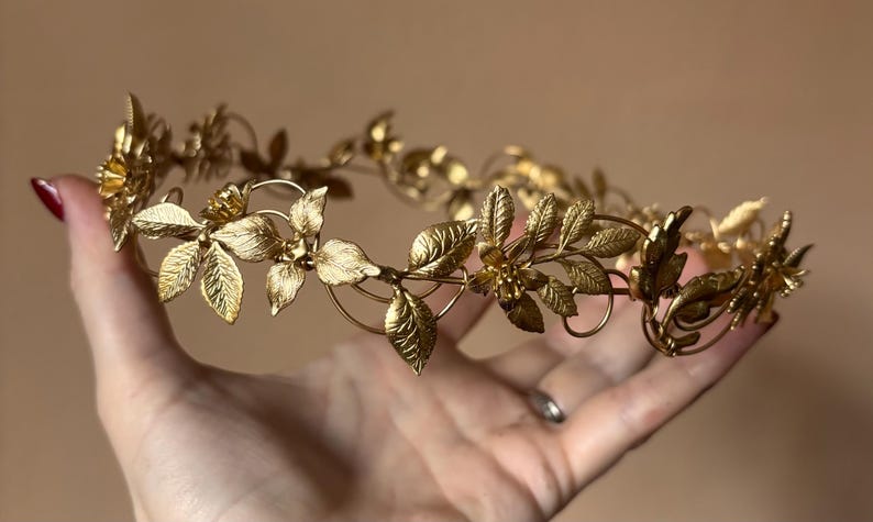 May include: A gold-toned metal crown featuring a delicate arrangement of textured leaves and small floral accents. The circular headpiece is constructed from thin, interconnected wire branches that form a continuous botanical pattern throughout.