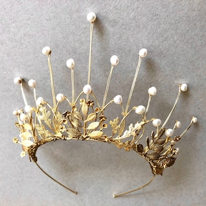 May include: A gold crown with a delicate design of leaves and pearls. The crown has a wire frame and is adorned with small white pearls.