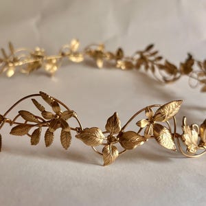 May include: A gold-toned metal headpiece featuring a delicate, continuous vine design. The accessory is adorned with intricate, textured leaves and small, five-petaled flowers arranged in a circular pattern, creating a flexible, lightweight crown.