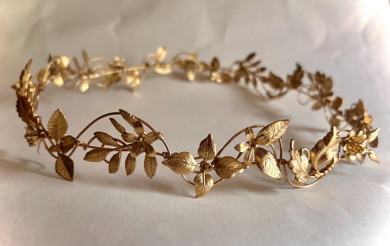 May include: A gold-toned metal headpiece featuring a delicate, continuous vine design. The accessory is adorned with intricate, textured leaves and small, five-petaled flowers arranged in a circular pattern, creating a flexible, lightweight crown.