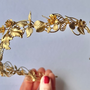 May include: A gold-colored metal floral crown with leaves and flowers. The crown is made of thin metal and has a delicate, intricate design. The leaves and flowers are detailed and add to the crown's elegant appearance. The crown is likely for a special occasion.