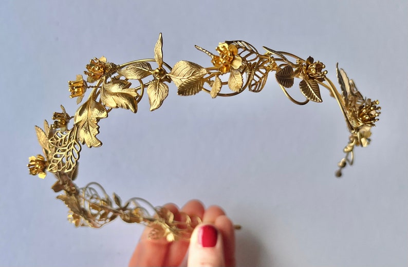 May include: A gold-colored metal floral crown with leaves and flowers. The crown is made of thin metal and has a delicate, intricate design. The leaves and flowers are detailed and add to the crown's elegant appearance. The crown is likely for a special occasion.