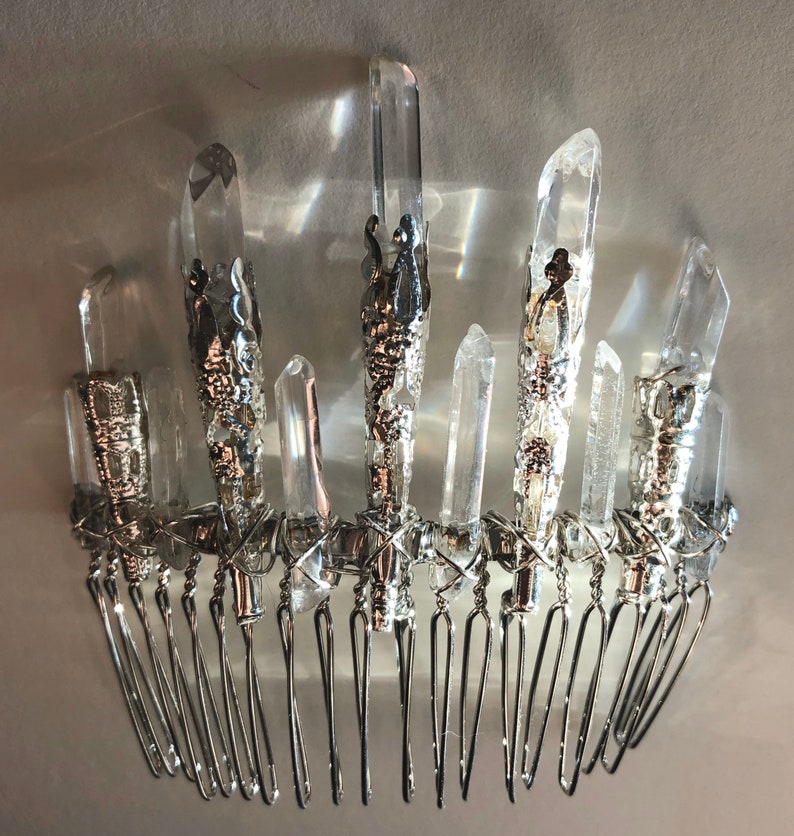 May include: A silver-toned hair comb adorned with clear quartz crystals. The crystals are of varying sizes and are set in ornate silver metalwork. The comb features multiple tines for securing in hair.