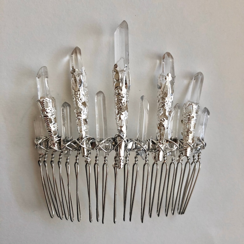 May include: A silver-toned hair comb adorned with clear quartz crystal points. The crystals are set in ornate silver metalwork and arranged above the comb's teeth. The comb is designed for hair styling and accessorizing.