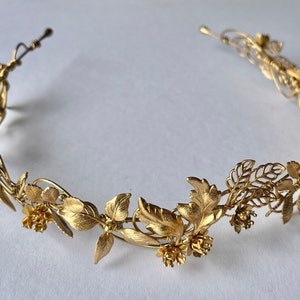 May include: A gold-colored metal floral crown featuring detailed leaves and small flowers. The crown is designed in a circular shape, suitable for weddings or special occasions. The metal has a matte finish.