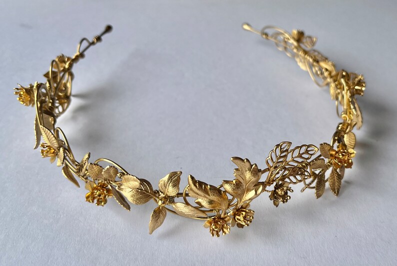 May include: A gold-colored metal floral crown featuring detailed leaves and small flowers. The crown is designed in a circular shape, suitable for weddings or special occasions. The metal has a matte finish.