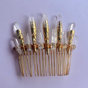 May include: A gold-colored hair comb adorned with clear quartz crystals. The comb features a series of gold-toned teeth, with the crystals arranged along the top. Some crystals are encased in gold filigree.