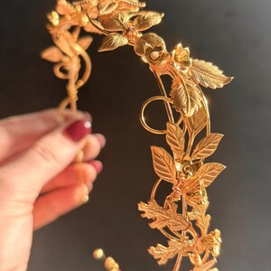 May include: A golden-colored floral crown with intricate leaf and flower details. The headpiece is held by a hand with red nail polish. The crown is designed with a delicate, curved structure, perfect for special occasions or themed events.
