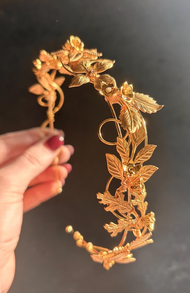May include: A golden-colored floral crown with intricate leaf and flower details. The headpiece is held by a hand with red nail polish. The crown is designed with a delicate, curved structure, perfect for special occasions or themed events.
