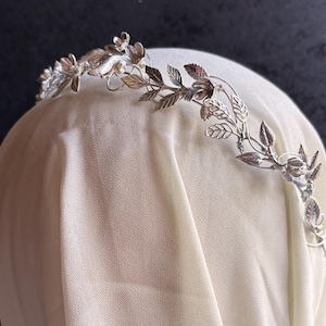 May include: A silver-colored floral crown with delicate leaves and flowers. The crown is designed with intricate details, suggesting a handcrafted or vintage style. The crown is likely intended for special occasions or as a bridal accessory.