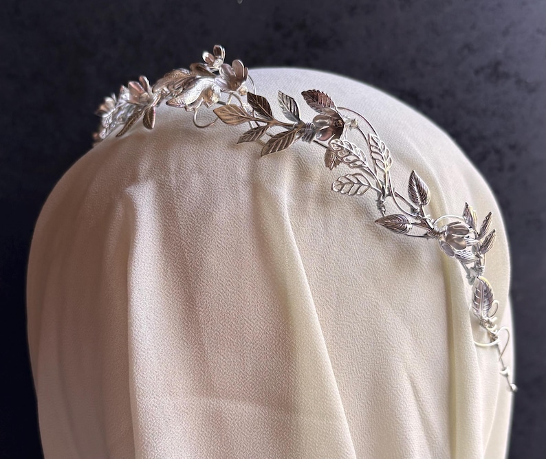 May include: A silver-colored floral crown with delicate leaves and flowers. The crown is designed with intricate details, suggesting a handcrafted or vintage style. The crown is likely intended for special occasions or as a bridal accessory.