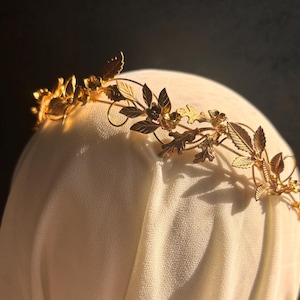 May include: A gold-colored metal crown with a floral and leaf design. The crown is placed on a white fabric surface, with the leaves and flowers intricately detailed. The crown's design includes various leaf shapes and small flower accents.