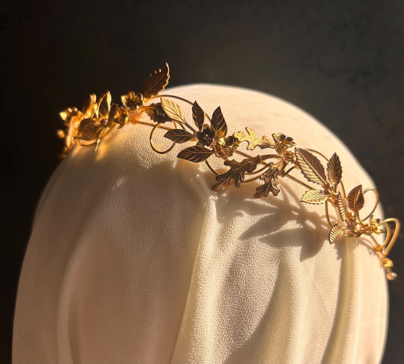 May include: A gold-colored metal crown with a floral and leaf design. The crown is placed on a white fabric surface, with the leaves and flowers intricately detailed. The crown's design includes various leaf shapes and small flower accents.