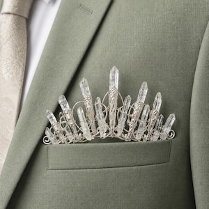 Pode incluir: Um acessório de bolso em fio prateado com múltiplos cristais de quartzo transparentes e pontiagudos dispostos em forma de coroa, inserido no bolso do peito de um casaco de fato verde sálvia com uma gravata clara estampada.