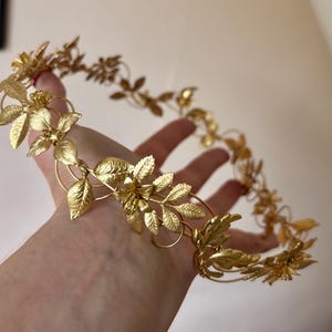 May include: A gold-colored metal crown featuring a continuous circular band adorned with intricate, textured leaf and small flower motifs. The metallic accessory has a polished finish and is constructed from thin, interconnected wire elements.