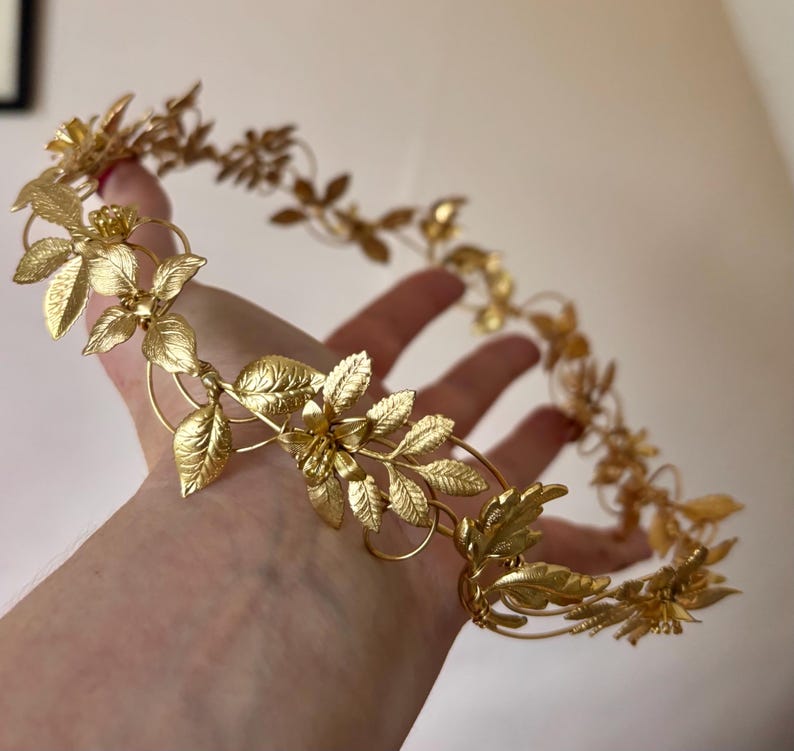 May include: A gold-colored metal crown featuring a continuous circular band adorned with intricate, textured leaf and small flower motifs. The metallic accessory has a polished finish and is constructed from thin, interconnected wire elements.