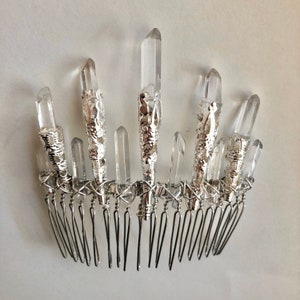 May include: A silver-toned hair comb adorned with clear quartz crystals. The crystals are of varying sizes and are set in ornate silver metal settings. The comb's teeth are thin and evenly spaced, creating a delicate and elegant accessory.