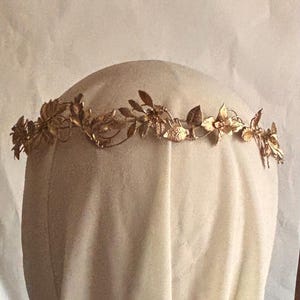 May include: A gold-colored metal headpiece featuring a delicate, continuous vine design with intricate leaves and small flower-like shapes. The accessory is constructed from thin, flexible wire elements that form a circular band for the head.