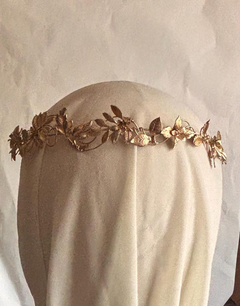 May include: A gold-colored metal headpiece featuring a delicate, continuous vine design with intricate leaves and small flower-like shapes. The accessory is constructed from thin, flexible wire elements that form a circular band for the head.