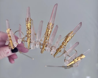 May include: A handcrafted crown featuring rose quartz crystals and gold-colored metal accents. The crown's design incorporates wire and decorative gold elements, with the crystals varying in size and shape. The overall aesthetic is elegant and ethereal.