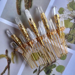 May include: A collection of gold-toned hair combs, each adorned with clear quartz crystals. The combs feature intricate gold filigree designs and are arranged on a floral background, creating an elegant and natural aesthetic.