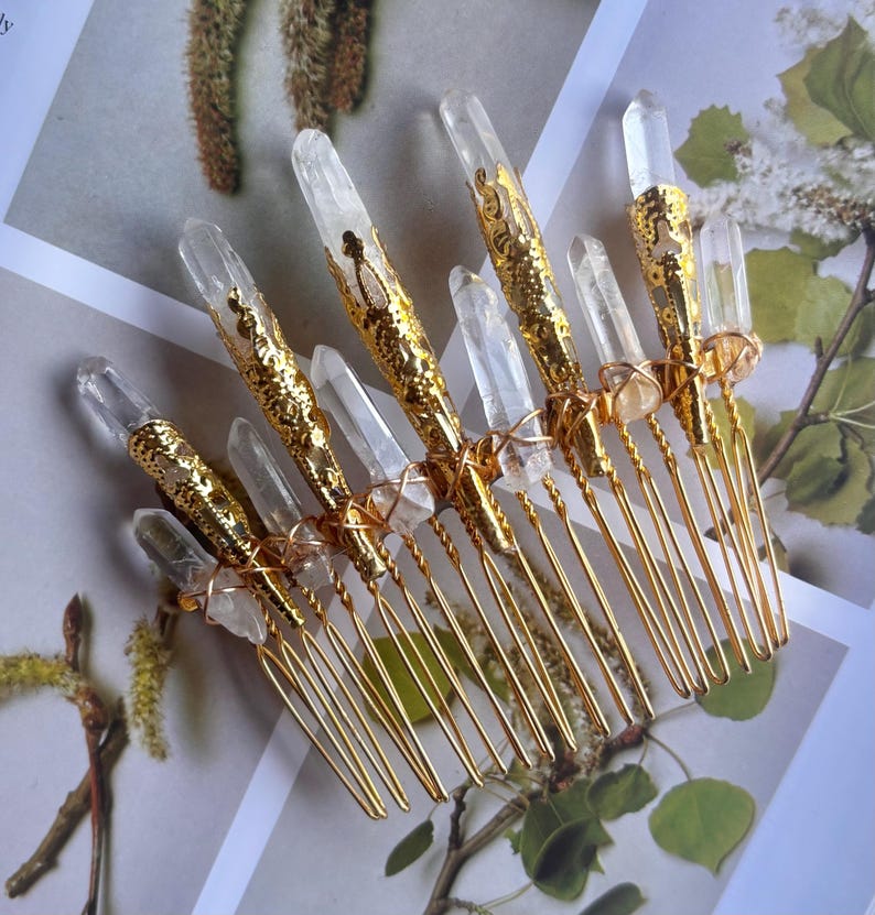 May include: A collection of gold-toned hair combs, each adorned with clear quartz crystals. The combs feature intricate gold filigree designs and are arranged on a floral background, creating an elegant and natural aesthetic.
