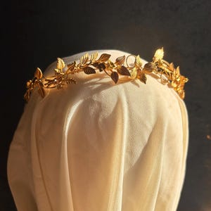 May include: A gold-colored leaf crown with intricate details. The crown features delicate leaves and floral accents, creating an elegant and natural design. The crown is displayed on a cream-colored fabric against a dark background, highlighting its golden hue.