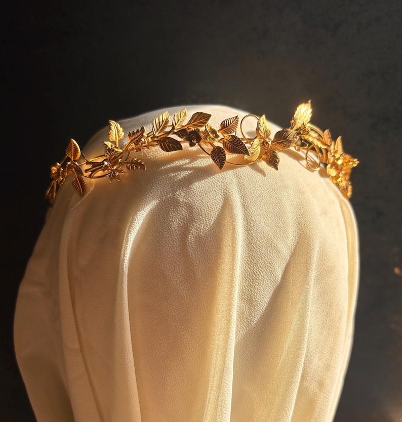 May include: A gold-colored leaf crown with intricate details. The crown features delicate leaves and floral accents, creating an elegant and natural design. The crown is displayed on a cream-colored fabric against a dark background, highlighting its golden hue.