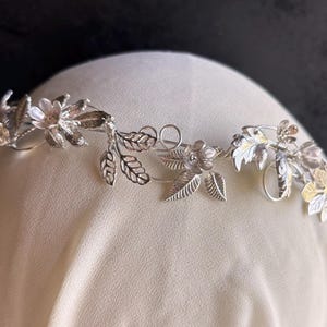 May include: A silver-toned floral crown with delicate leaves and flowers. The crown is designed with intricate details, featuring small blossoms and detailed leaf patterns. The headpiece is displayed on a cream-colored surface against a dark background.