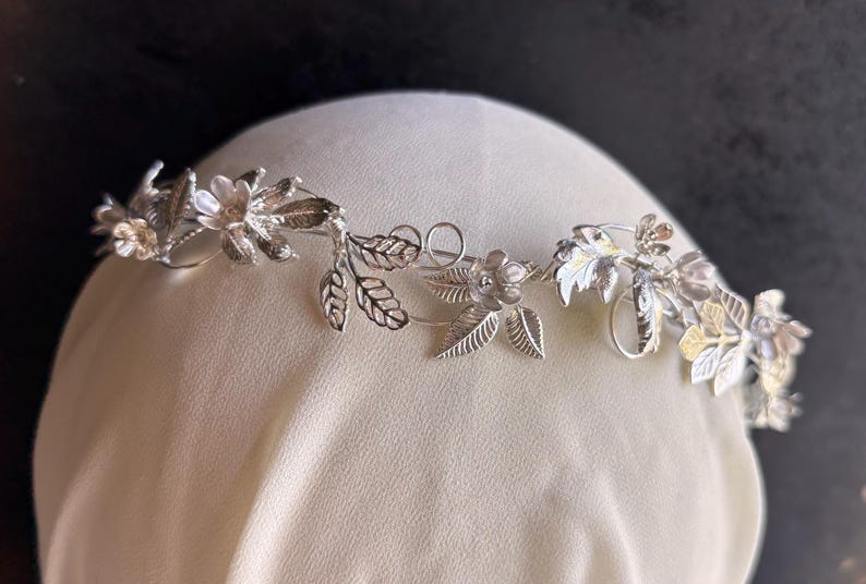 May include: A silver-toned floral crown with delicate leaves and flowers. The crown is designed with intricate details, featuring small blossoms and detailed leaf patterns. The headpiece is displayed on a cream-colored surface against a dark background.