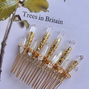 May include: A gold-colored hair comb with clear quartz crystals and decorative gold accents. The comb features multiple tines and is adorned with several clear quartz crystals. The text "Trees in Britain" is visible in the background.