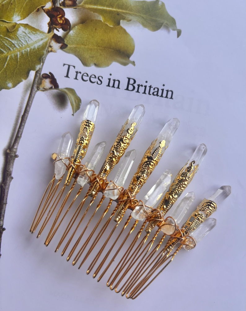 May include: A gold-colored hair comb with clear quartz crystals and decorative gold accents. The comb features multiple tines and is adorned with several clear quartz crystals. The text "Trees in Britain" is visible in the background.
