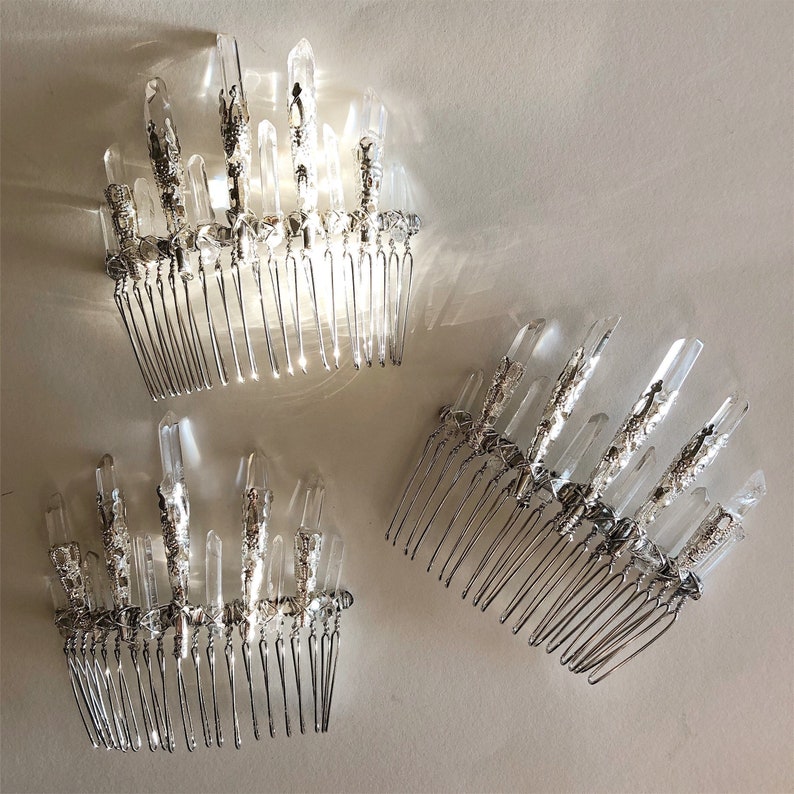 May include: Three silver hair combs adorned with clear quartz crystals. The crystals are arranged in a vertical, spiky pattern, and the combs have a delicate, intricate design. The combs are displayed on a light-colored surface.