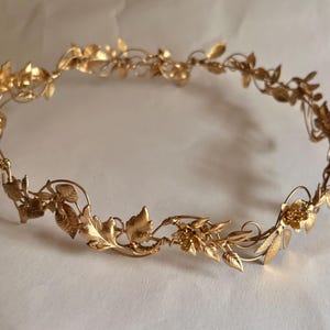 May include: A gold-colored metal crown featuring a continuous vine design. The circular headpiece is adorned with intricate, textured leaves and small floral accents connected by thin, flexible wire branches that form a delicate, open structure.
