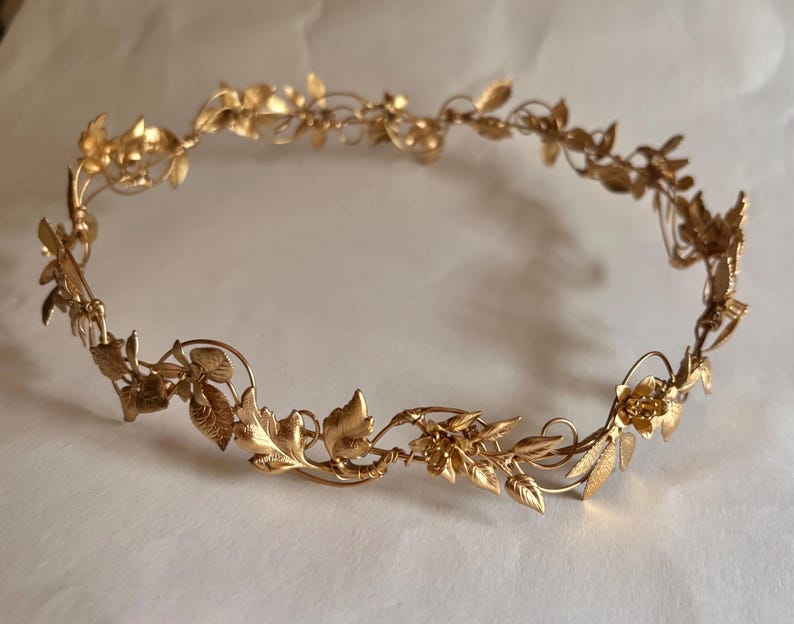 May include: A gold-colored metal crown featuring a continuous vine design. The circular headpiece is adorned with intricate, textured leaves and small floral accents connected by thin, flexible wire branches that form a delicate, open structure.
