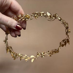 May include: A gold-toned metal headpiece featuring a continuous vine design with intricate leaves and small flower blossoms. The circular accessory is constructed from thin, flexible wire branches that form a delicate, botanical-inspired crown.