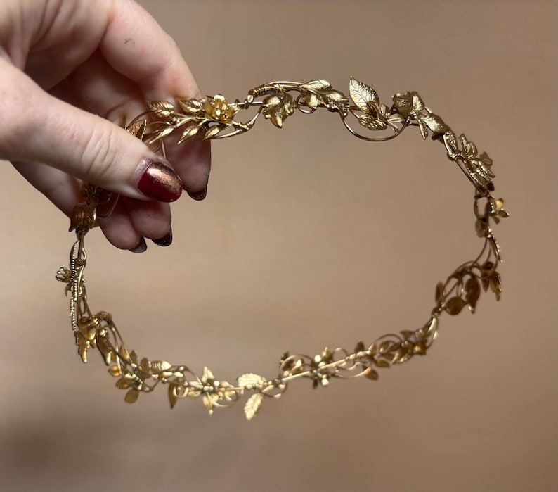 May include: A gold-toned metal headpiece featuring a continuous vine design with intricate leaves and small flower blossoms. The circular accessory is constructed from thin, flexible wire branches that form a delicate, botanical-inspired crown.
