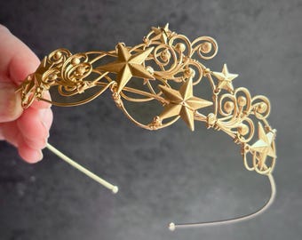 May include: A gold-colored star-themed crown. The crown features multiple five-pointed stars and decorative swirls. The headband is held by a hand against a dark background. The crown is made of metal.