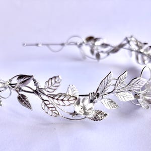 May include: A silver-colored leaf crown, featuring detailed leaf and floral designs. The crown is made of a shiny metal and is designed to be worn on the head. The leaves and flowers are intricately crafted, creating an elegant and delicate appearance.
