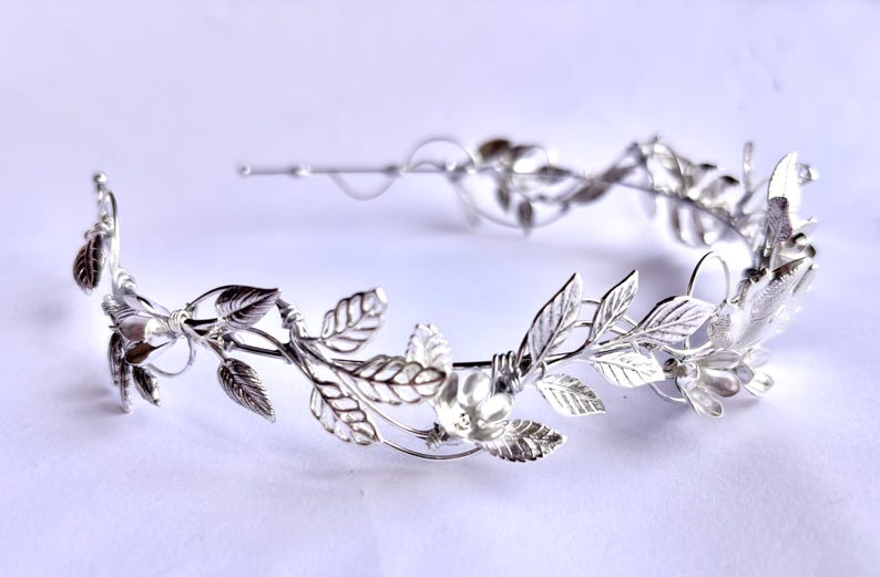 May include: A silver-colored leaf crown, featuring detailed leaf and floral designs. The crown is made of a shiny metal and is designed to be worn on the head. The leaves and flowers are intricately crafted, creating an elegant and delicate appearance.