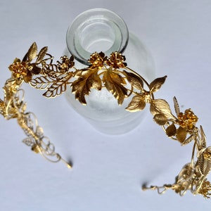 May include: A gold-colored metal floral crown with leaves and flowers. The crown is designed with intricate details, including delicate leaves and small, clustered flowers. The metal has a shiny finish, reflecting light. The crown is likely for a special occasion.