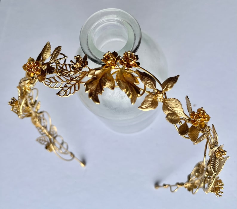 May include: A gold-colored metal floral crown with leaves and flowers. The crown is designed with intricate details, including delicate leaves and small, clustered flowers. The metal has a shiny finish, reflecting light. The crown is likely for a special occasion.