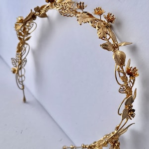 May include: A gold-colored metal headband with floral and leaf details. The decorative crown features intricate designs of flowers and leaves, creating an elegant and ornate accessory. The headband is designed to be worn on the head.