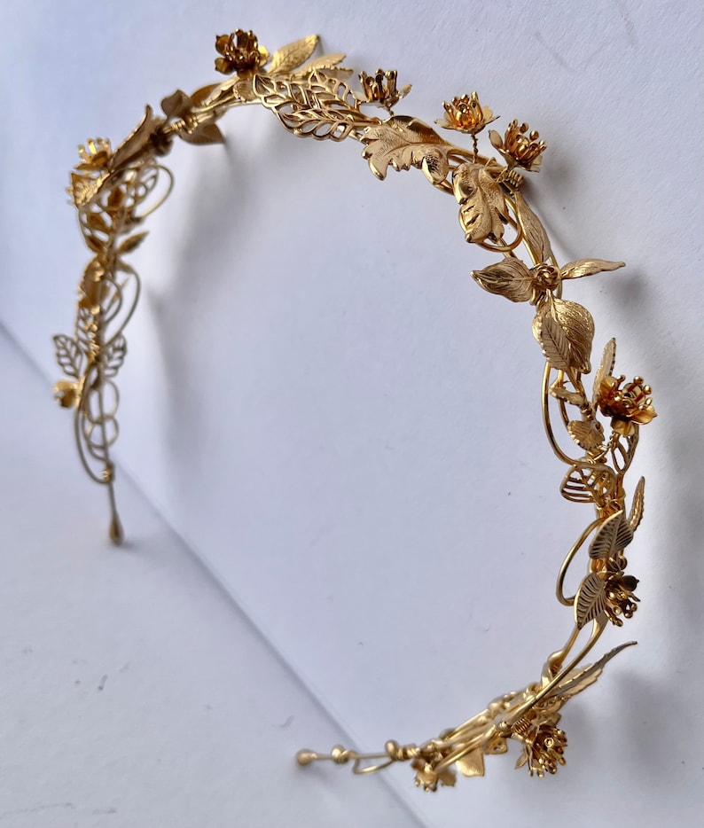 May include: A gold-colored metal headband with floral and leaf details. The decorative crown features intricate designs of flowers and leaves, creating an elegant and ornate accessory. The headband is designed to be worn on the head.