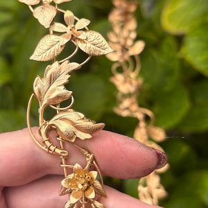 May include: A gold-toned metal headpiece featuring a continuous vine design with intricate leaves and small flowers. The accessory is constructed from thin, flexible wire branches that connect the botanical elements in a repeating pattern.