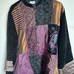May include: A patchwork jacket in a variety of textures and colours. The jacket features black velvet sleeves, a black velvet collar, and a mix of fabrics in shades of purple, gold, and patterned textiles. It has wooden buttons and embroidered details.