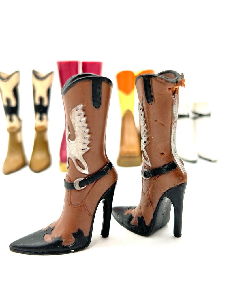 May include: A pair of brown and black high-heeled cowboy boots with silver accents. The boots have pointed toes and a black strap around the ankle. Other boots in various colors are visible in the background.