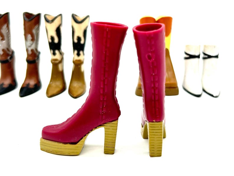 May include: A collection of miniature boots in various styles and colors. The foreground features a pair of bright pink platform boots with gold heels. Other boots include brown, white, and black cowboy boots, and a pair of white ankle boots.