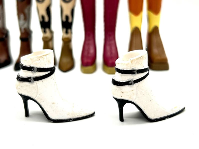 May include: Two white ankle boots with black straps and buckles, set against a blurred background of various colored cowboy boots. The boots have pointed toes and black stiletto heels.