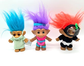 May include: Three Troll dolls with colorful hair and outfits. One has blue hair and a face mask, another has purple hair and a pink swimsuit, and the third has red hair and a black outfit with a diving mask.
